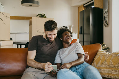 Happy multiracial couple enjoying sharing smart phone while sitting on sofa in living room at home