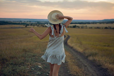 Woman in the hat and dress is on the field at sunset on the track