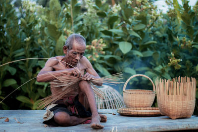 Full length of shirtless man making wicker basket 