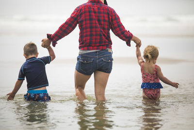 Rear view of mother helping kids wade through water at the beach.