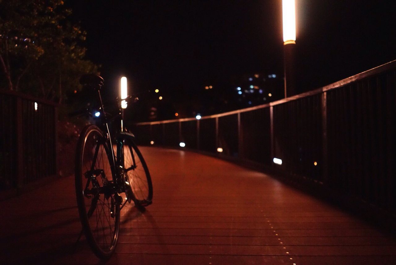 night, illuminated, transportation, street light, bicycle, lighting equipment, the way forward, built structure, building exterior, street, city, architecture, mode of transport, land vehicle, light - natural phenomenon, road, outdoors, sidewalk, parking, railing