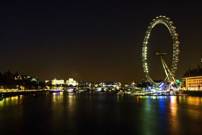 Illuminated ferris wheel by river against sky in city at night