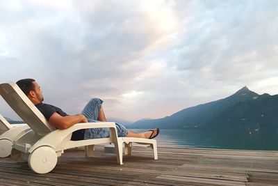 Man reclining on lounge chair by lake against mountain