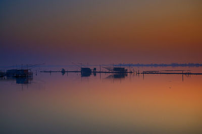 Fishing huts by stilts on sea against sky during sunset