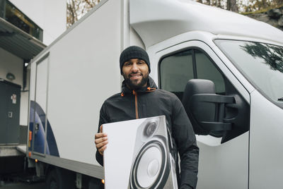 Portrait of smiling male worker holding box while standing against delivery van