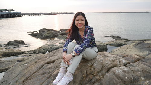 Portrait of smiling young woman sitting on rock by sea