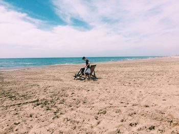 Side view of teenage boy standing by deck chairs at beach against cloudy sky