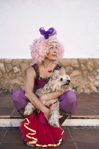 Full body of mature female clown in funny pink wig and costume embracing adorable fluffy dog and looking away while sitting on street after performance