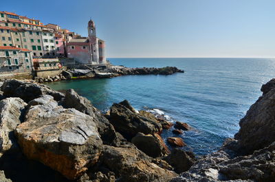 Scenic view of rocks at seashore by city against clear sky