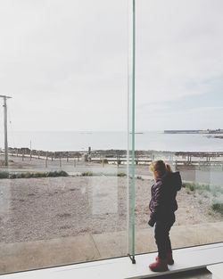 Full length of girl standing on window sill looking at sea against sky