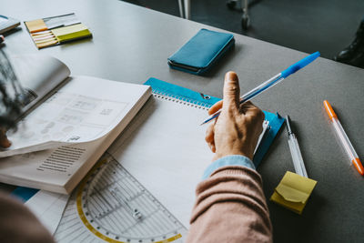 Cropped image of mature female student sitting with book and pen at desk in classroom