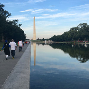 People walking on walkway by canal at washington monument against blue sky