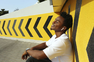 Smiling man with eyes closed listening to music sitting near wall