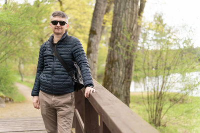 Middle-aged man in sunglasses and thin down jacket on wooden footbridge near lake in early spring