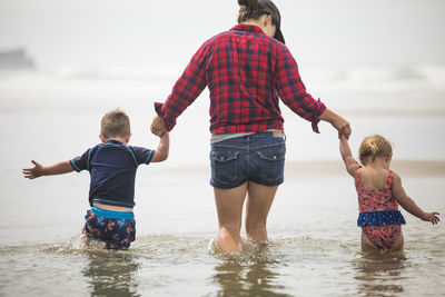 Rear view of mother helping kids wade through water at the beach.