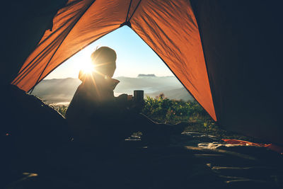 Silhouette man with mug in tent on mountain against bright sun in sky during sunrise