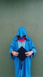 Man wearing blue graduation gown holding black mortarboard while standing against wall