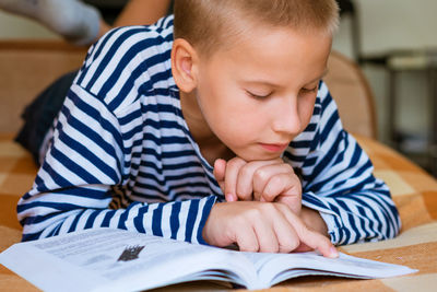 A cute school-age boy reads a book while lying on his stomach on the couch does