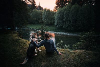 Rear view of woman sitting with dog at riverbank in forest