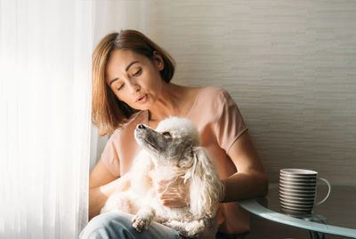 Single young brunette woman with poodle dog on hands sitting by the window at home