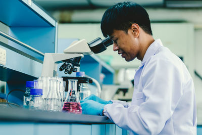 Asian man lab technician in protective glasses and gloves sits next to a microscope in laboratory.