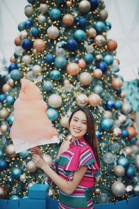 Portrait of smiling young woman standing by christmas tree holding cotton candy outdoors