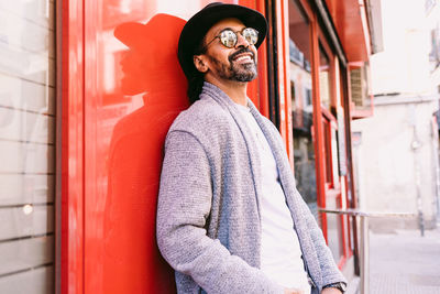 Happy hispanic male in stylish clothes with sunglasses and hat smiling and looking away while leaning on red wall of building on sunny day on city street