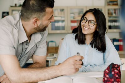 Smiling male teacher explaining student while leaning on table in classroom