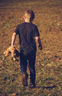 Rear view of boy walking with stuffed toy on field