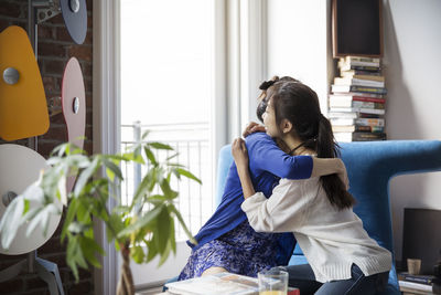 Mother embracing daughter sitting on sofa at home