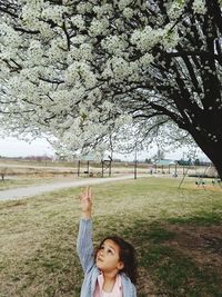 Cute girl standing with raised hand below flowers on tree