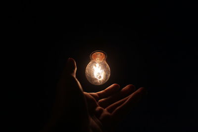 Close-up of hand by illuminated light bulb over black background