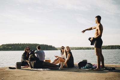 Shirtless man pointing while talking to friends on jetty at lake against sky