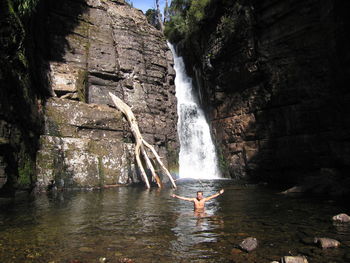 Carefree shirtless man with arms outstretched swimming in river against waterfall and rock formation