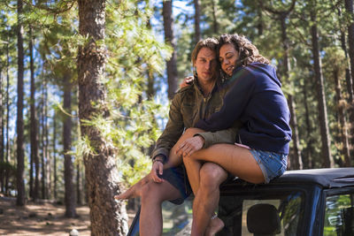 Young couple sitting on car roof against trees in forest