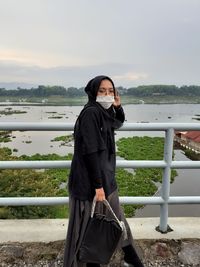 Woman standing by railing against lake against sky