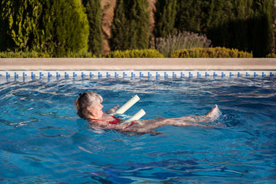 Side view of cheerful elderly female with gray hair swimming in pool with aqua noodle