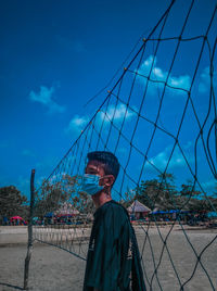 Portrait of man standing by fence against blue sky