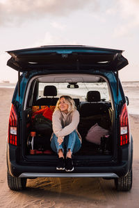 Full length of young woman looking away while sitting on car trunk at beach against sky