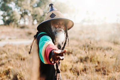 Cheerful old ethnic rastafari with dreadlocks looking at the camera in standing in a dry meadow in the nature