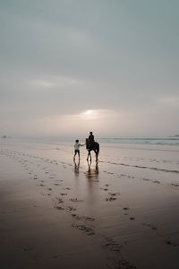 Silhouette woman walking at beach against sky during sunset and a person riding a horse