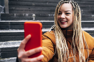 Young smiling woman taking selfie through mobile phone while sitting on steps