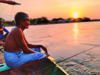 Side view of young man sitting against sky during sunset