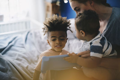 Smiling boy looking at mother with toddler teaching digital tablet at home