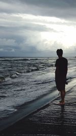 Rear view of man standing on beach against cloudy sky