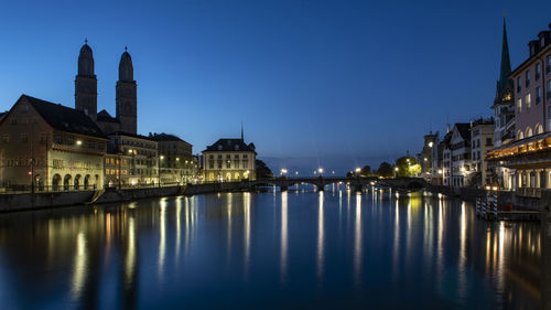 Illuminated buildings by river against blue sky