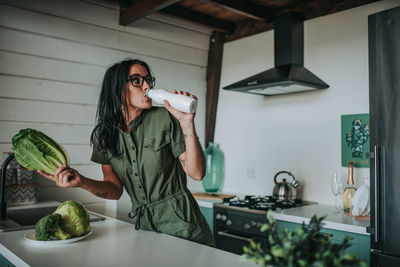 Smiling woman drinking milk and eating vegetables in kitchen at home