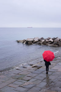 Woman carrying red umbrella standing at sea shore