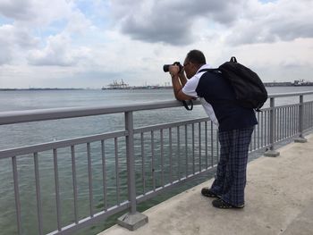Full length of man photographing through camera while standing on bridge over sea