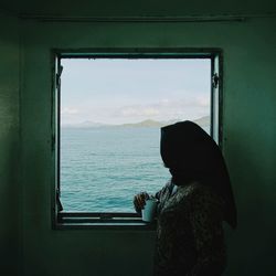 Side view of woman in hijab having drink while looking at sea through window